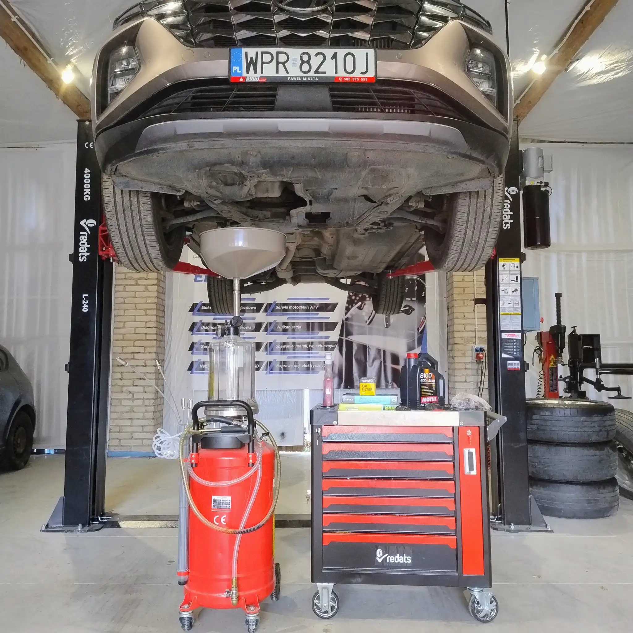 Samochód SUV podniesiony na podnośniku dwukolumnowym, z wózkiem serwisowym i urządzeniem do wymiany oleju.